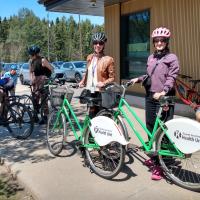 two women with helmets and sunglasses stand beside TBDHU branded bicycles